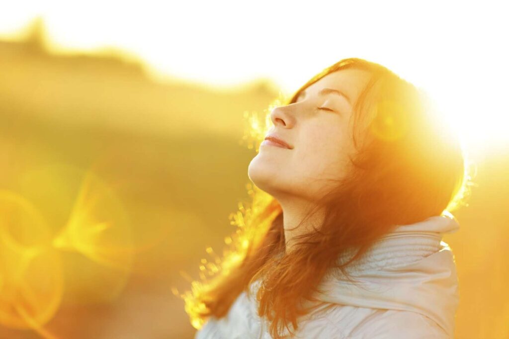 A person with long hair stands outdoors, eyes closed, soaking in the sun. Clad in a light-colored jacket, they exude a serene glow reminiscent of a plant-based lifestyle, as warm sunlight casts lens flares across the vibrant backdrop.
