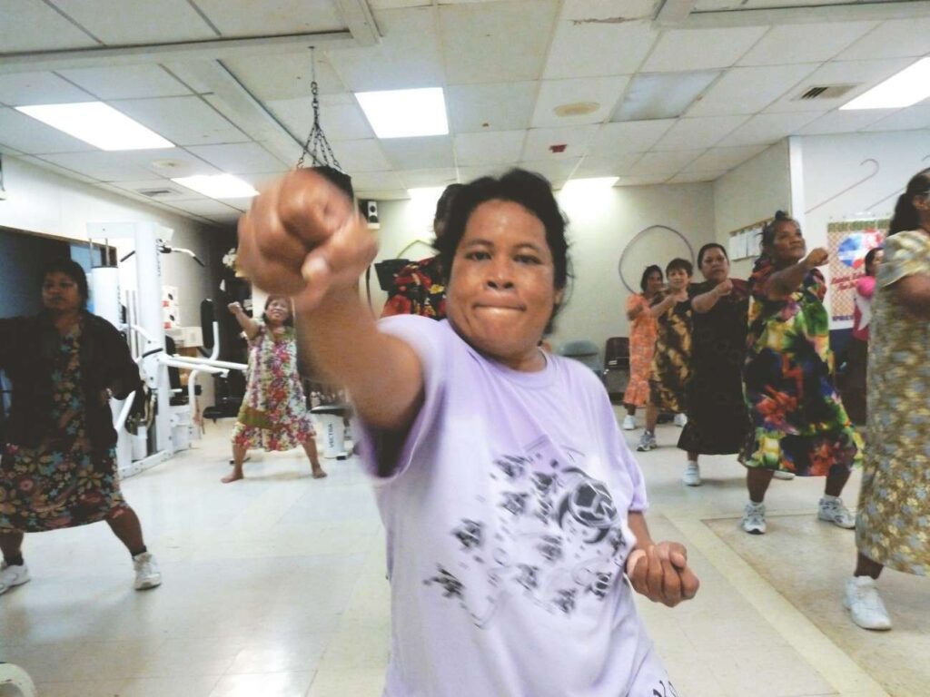 A group of women in colorful dresses, guided by a dietitian, participate in an indoor exercise class. The woman in the foreground extends her fist toward the camera. With fluorescent lighting and exercise equipment around, everyone appears focused and engaged on their wellness journey.