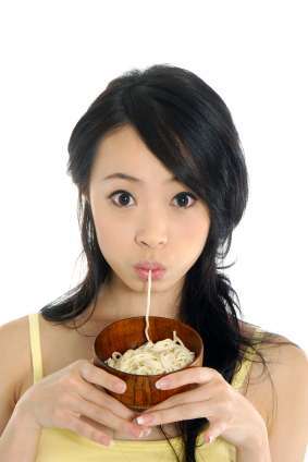 A person with long dark hair is enjoying plant-based noodles from a wooden bowl. They are wearing a yellow top and have a surprised expression, with noodles stretching from the bowl to their mouth. The background is plain white.