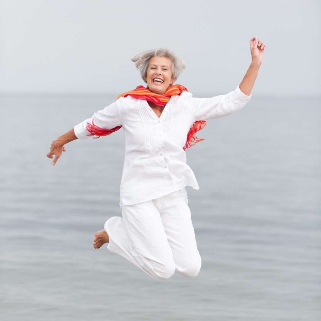 An elderly woman, embracing a vibrant vegetarian lifestyle, joyfully jumps in the air on a beach, wearing a white outfit and red scarf with the sea stretching behind her.