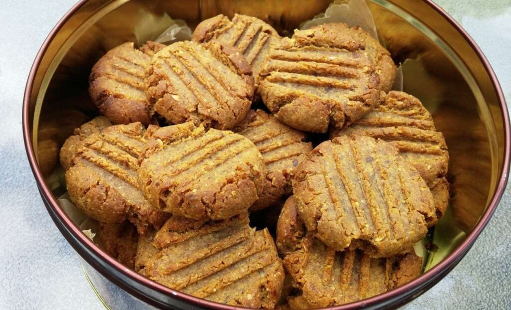 A bowl filled with golden-brown, plant-based cookies, each with a crisscross pattern on top. The cookies appear rustic and crumbly, stacked neatly in a circular metal container.