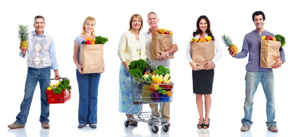 Five people standing with a fresh variety of plant-based fruits and vegetables. Three hold paper bags, one carries a basket, and another proudly displays a pineapple. One person pushes a shopping cart filled with produce. They are all smiling against a plain white background, embodying the joy of vegetarian living.