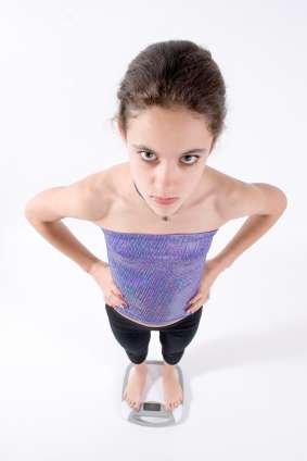 A person with brown hair stands on a digital scale, embodying confidence. They are looking directly up at the camera with hands on their hips, wearing a purple top and black pants. As someone who consults a dietitian for their plant-based journey, they radiate commitment to wellness amidst the white backdrop.