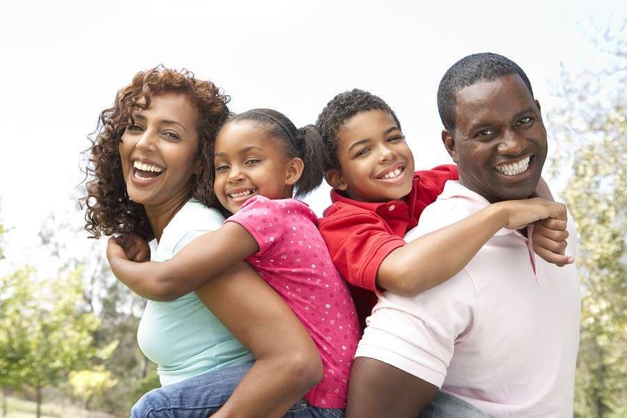A cheerful family enjoys a sunny afternoon in the park. The parents, both smiling vegan enthusiasts, are giving piggyback rides to their joyful children. The mother and daughter are on the left, while the father and son are on the right, all beaming against a lush green backdrop.
