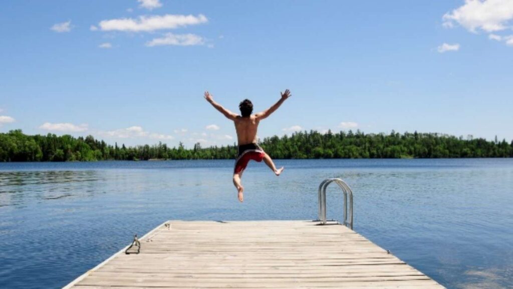A person in red shorts, perhaps a vegan adventurer, leaps off a wooden dock into a calm lake. The clear sky hosts a few clouds, while a line of trees paints the background—a scene as refreshing as living plant-based.