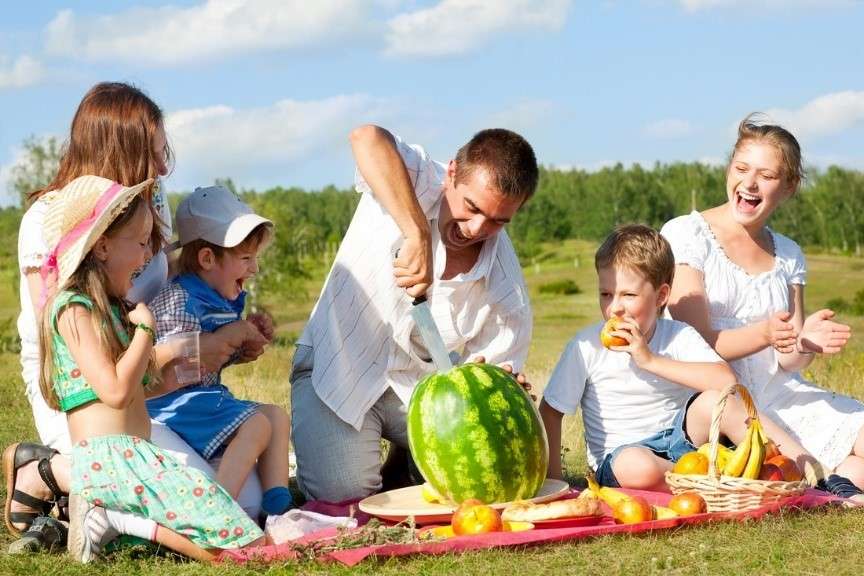 A group of six people, including children and adults, are having a plant-based picnic on the grass. A man in the center is cutting a watermelon while others watch and smile. A basket of fresh fruit is nearby. Its a sunny day with a blue sky and scattered clouds.