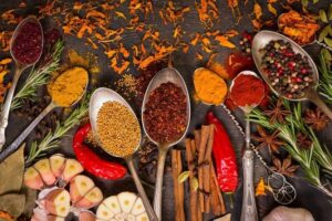 Spoons filled with vibrant spices like turmeric, chili powder, mustard seeds, and peppercorns are artfully arranged on a dark surface. Surrounding them are star anise, cinnamon sticks, garlic, red chili peppers, and rosemary—a feast for any vegan or plant-based diet enthusiast.