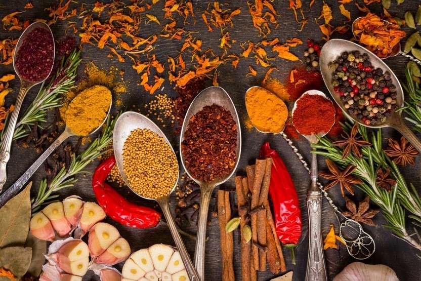 Spoons filled with vibrant spices like turmeric, chili powder, mustard seeds, and peppercorns are artfully arranged on a dark surface. Surrounding them are star anise, cinnamon sticks, garlic, red chili peppers, and rosemary—a feast for any vegan or plant-based diet enthusiast.