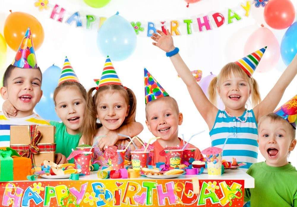 Six children wearing colorful party hats are celebrating a birthday with a delightful spread of plant-based snacks and drinks. They sit around a table filled with presents, under a Happy Birthday banner and balloons. The children are smiling and joyful as they enjoy the vegetarian feast.