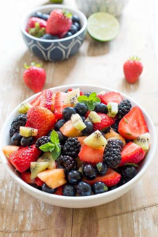 A white bowl filled with sliced strawberries, blueberries, blackberries, and kiwi, garnished with mint leaves. This vibrant plant-based spread showcases a dietitians dream of fresh fruits. Two whole strawberries and a sliced lime rest on a wooden surface. A patterned bowl partially visible behind.