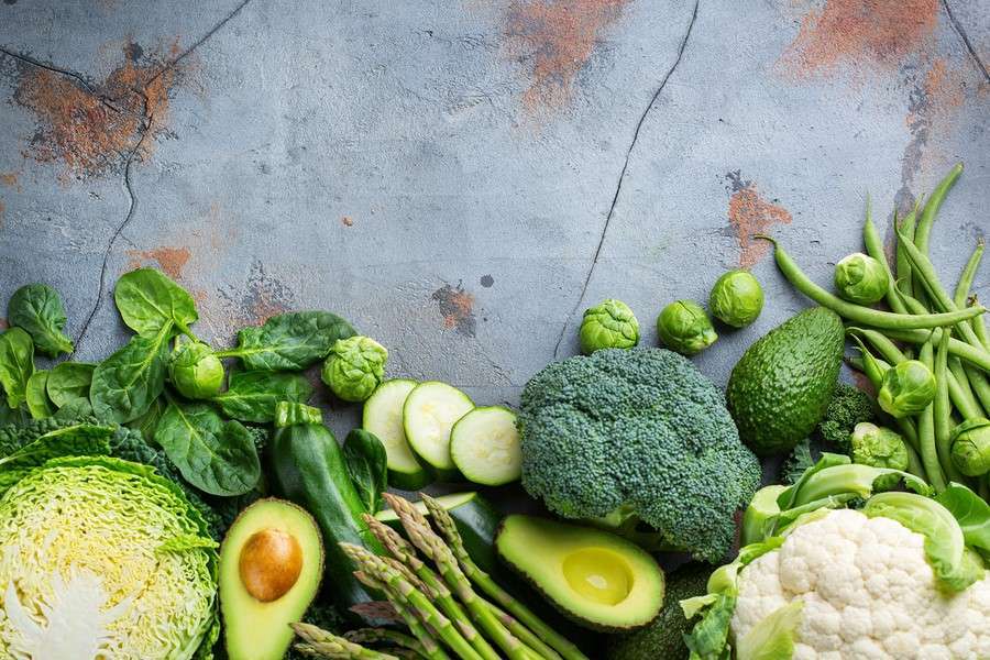 A variety of green vegetables, including spinach, Brussels sprouts, zucchini, broccoli, asparagus, avocado, cabbage, green beans, and cauliflower—all essential in a plant-based diet—are arranged on a rustic, textured gray surface.