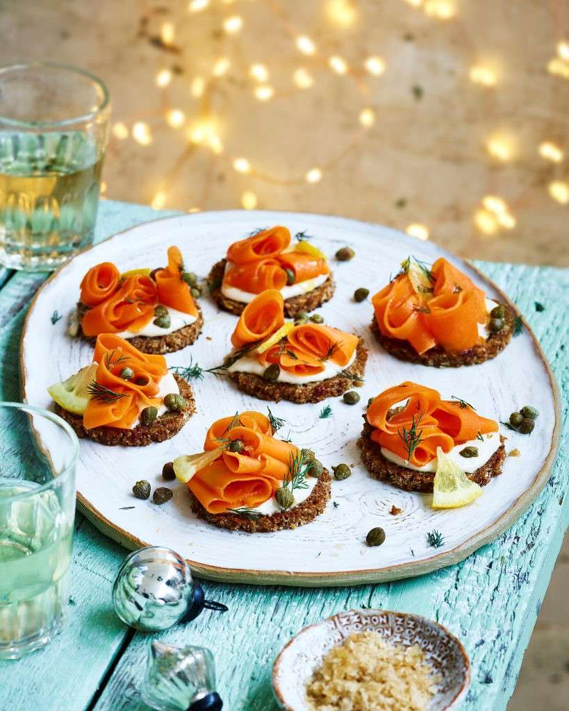 A white plate on a rustic table holds six vegetarian appetizers with rye bread, topped with cream, thin carrot ribbons, capers, and dill. A small dish of brown sugar and glasses with light drinks are nearby. Soft, warm, blurred lights in the background add ambiance.