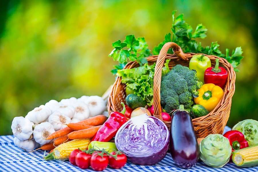A wicker basket filled with broccoli, red and yellow bell peppers, and cucumbers. Surrounding it are garlic bulbs, carrots, cherry tomatoes, corn, eggplant, red cabbage, and leafy greens—a vibrant spread perfect for any vegetarian feast on a blue and white checkered tablecloth.
