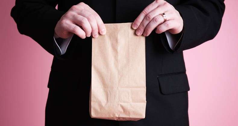 A person wearing a black suit is holding a small, closed brown paper bag with both hands against a pink background. Perhaps it contains a vegan treat curated by an expert dietitian. Only the hands and part of the suit are visible.