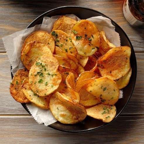 A bowl of homemade potato chips, perfect for a plant-based snack, is garnished with chopped parsley and set on parchment paper. The chips are golden brown and slightly crispy, resting invitingly on a wooden table.