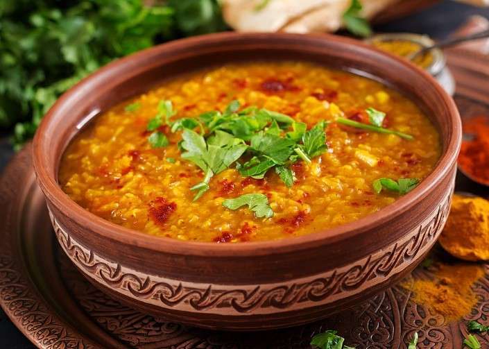 Ceramic bowl filled with vibrant yellow lentil soup, garnished with fresh cilantro and a drizzle of red oil. Perfect for a vegan spread, the intricately patterned bowl rests on a decorative platter, surrounded by greenery and spices.