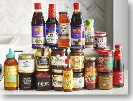 A variety of condiment bottles and jars, including plant-based soy sauce, hoisin sauce, pesto, peanut butter, chili sauce, and spreads, are arranged in rows on a white surface. Different shapes and sizes represent various brands.