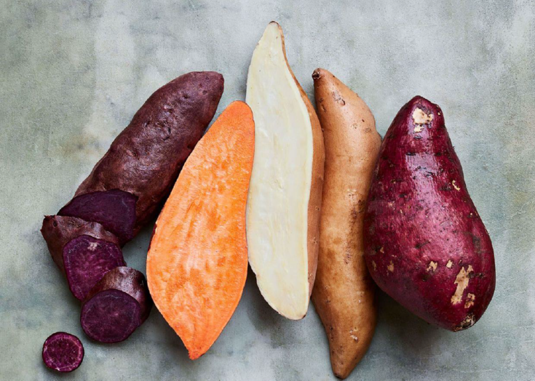 Various types of sweet potatoes and yams are displayed on a textured surface. The image shows purple, orange, and pale yellow varieties, both whole and sliced, perfect for any vegetarian or plant-based diet, highlighting the vibrant color contrast and variety in texture.