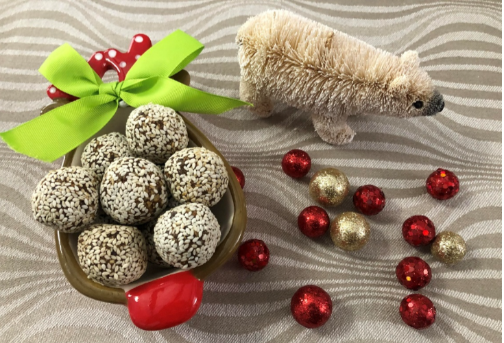 A ceramic reindeer-shaped dish filled with vegan sesame-covered balls, tied with a green bow. Beside it, a small plush polar bear and scattered red and gold glittery balls lie on a wavy patterned surface.
