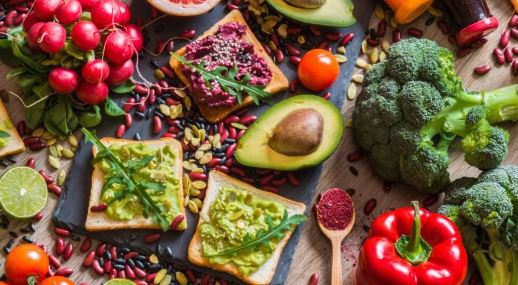 A colorful spread featuring vegan avocado toasts with arugula, halved avocados, broccoli, radishes, cherry tomatoes, a lime, and a bell pepper. Seeds and grains are scattered around, creating a vibrant and fresh atmosphere worthy of any dietitians recommendation.