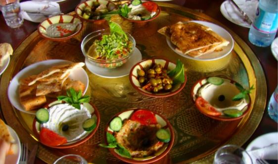 A variety of Middle Eastern dishes, perfect for a vegetarian feast, are arranged on a large, ornate brass platter. Dishes include hummus, flatbread, salads, and dips with garnishes like cucumber slices, tomatoes, and olives. Bottled water is visible in the background.