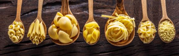 Seven wooden spoons rest on a wooden surface, each cradling a type of uncooked pasta—elbow macaroni, penne, conchiglie, farfalle, tagliatelle, fusilli, and orzo—all perfect for crafting delicious vegetarian dishes.