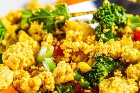 Close-up of a vibrant vegan tofu scramble mixed with chopped kale, red and green bell peppers, and spring onions. The dish appears well-seasoned, with a golden hue, suggesting turmeric or curry powder seasoning. A fork is partially visible in the background.