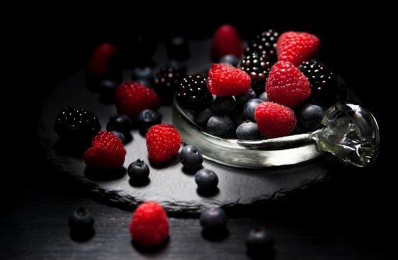 A glass bowl overflowing with plant-based delights like raspberries, blackberries, and blueberries rests on a dark slate surface. Additional berries are scattered around it, set against a dark background with dramatic lighting highlighting the vibrant red and deep blues.