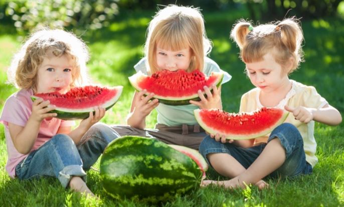 Three young children sit on the grass, enjoying large slices of watermelon on a sunny day. A whole watermelon, perfect for a plant-based snack, sits in front of them. They are smiling and appear to be having fun outdoors.