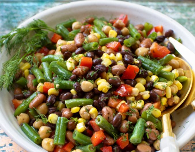 A vibrant, plant-based bean salad in a white bowl, featuring green beans, black beans, kidney beans, chickpeas, corn, and diced red bell peppers. Fresh dill is sprinkled on top with a golden spoon beside it for a perfect vegan delight.